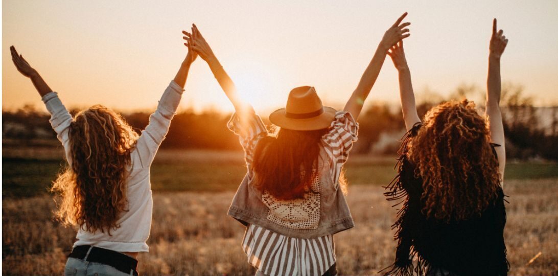 3 jeunes filles de dos qui lèvent les bras devant un coucher de soleil